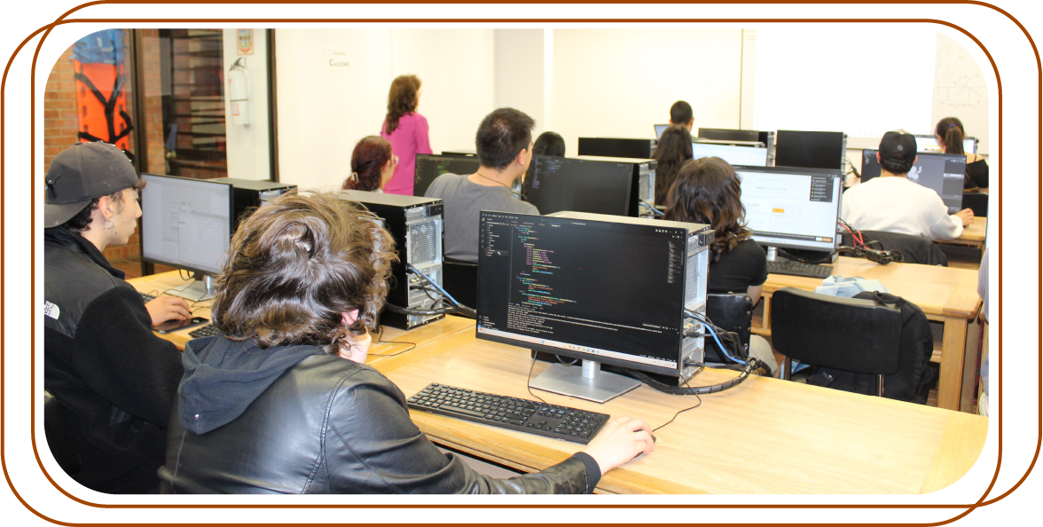 aula con varias personas trabajando en computadoras de escritorio. Los monitores exhiben código de programación y otras interfaces de software.