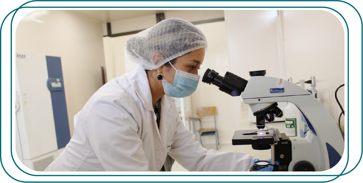 na persona en un entorno de laboratorio. Está vestida con una bata de laboratorio y una red para el cabello, y está utilizando un microscopio.