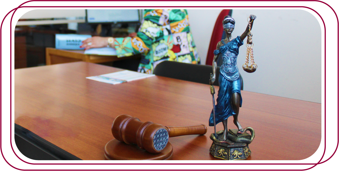 una figura de la Justicia,jJunto a la figura hay un martillo de madera sobre una superficie de mesa. En el fondo, se ve a una persona en una oficina.
