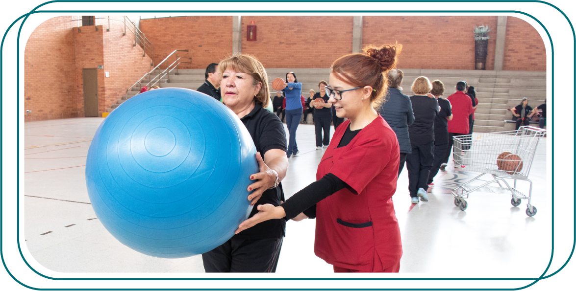 dos personas con los rostros pixelados, una de ellas sosteniendo una gran pelota de ejercicio de color azul, en una instalación deportiva cubierta, ya que hay otras personas en el fondo que parecen estar realizando actividades físicas, en una sesión de terapia física.