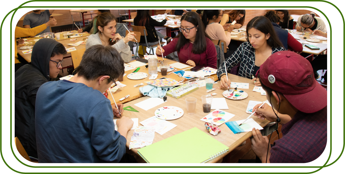 grupo de personas jóvenes reunidas alrededor de una mesa realizando actividades de pintura y arte. Cada persona está enfocada en su propio proyecto.